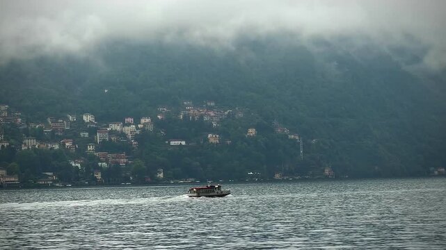 Lake Como, Moltrasio, Italy. Boat moving smoothly on a calm lake surrounded by misty mountains and vibrant houses along the shoreline