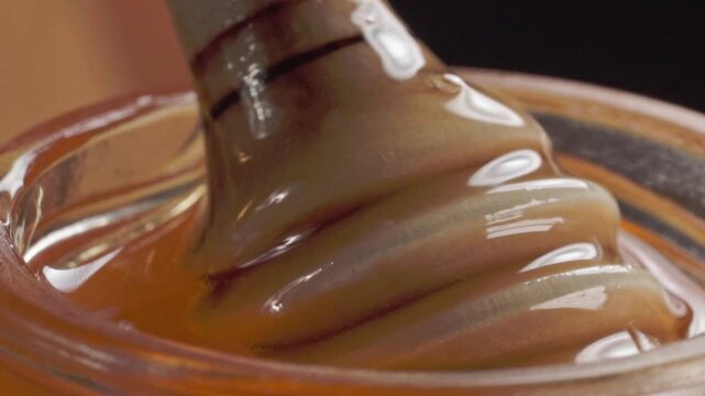 Macro close up of thick amber colored syrup with a glossy texture being stirred in a glass container, showing smooth folds and viscous consistency