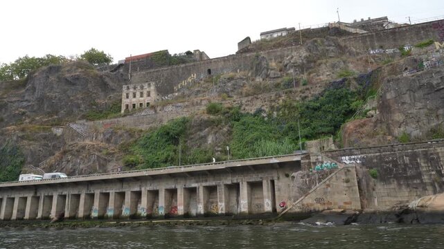 Rugged cliffside meets water's edge, ancient stone pier extends. Weathered walls, greenery, and distant buildings frame the scene. , quiet, solitary, urban decay blends with nature