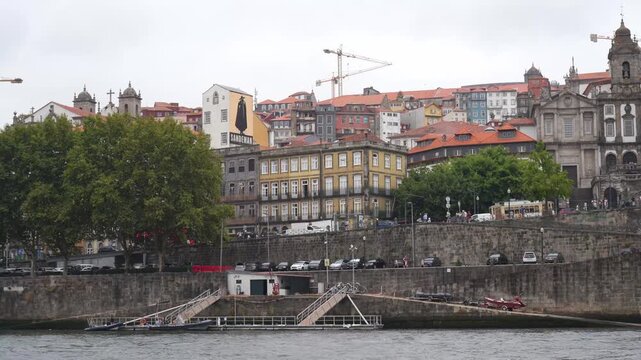 Overcast sky hovers above historic port cityscape. Red-tiled roofs cascade up stone cliffs beside calm river. Trees line waterfront with gentle breeze rustling leaves