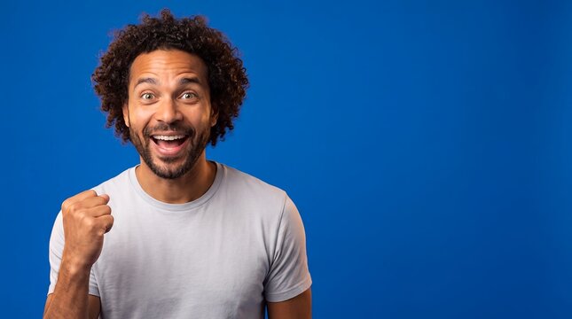 Adult man delighted surprise raised fist blue solid background portrait An adult man raises a clenched fist with an excited open smile for winning moments and personal success campaigns