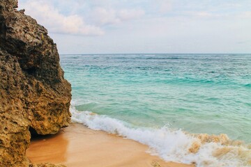 Wide shot of turquoise sea water breaking into soft white foam along golden sand shore next to a large coral rock cliff at Geger Beach, Bali, Indonesia. Travel theme. © Arlina Elsiska