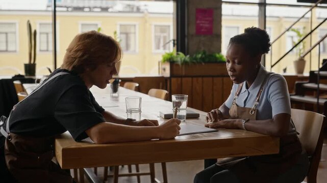 Diverse African American woman and Caucasian man in aprons discussing documents at cafe table. Footage suitable for small business management, hospitality training, and teamwork projects