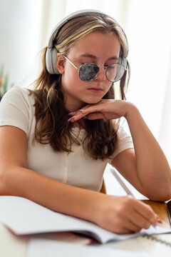 A teenage girl with strabismus wearing special corrective eyeglasses with a lens patch studies at a desk. She wears headphones while writing in a notebook during school preparation.