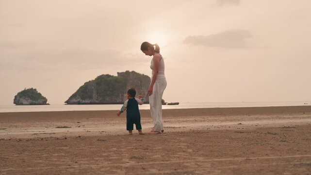 4K Asian mother holding hands with toddler son walking on sandy beach together with sunset time.
