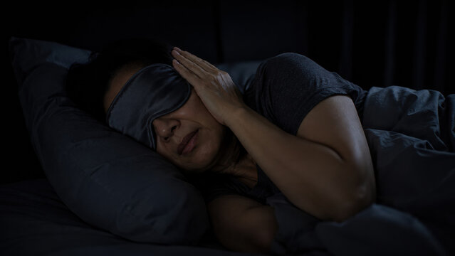 A woman lying in bed wearing a soft eye mask in a dark room, one hand resting against their temple over the mask. Migraine dark-room retreat concept.