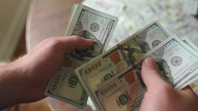 Caucasian man organizes and counts multiple stacks of hundred-dollar bills on a wooden table with more cash visible in the background