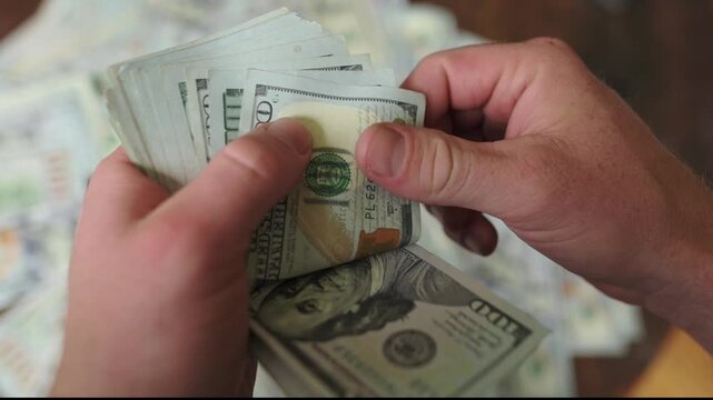 Hands of a person methodically counting and organizing hundred-dollar bills on a wooden table, surrounded by additional cash in the background