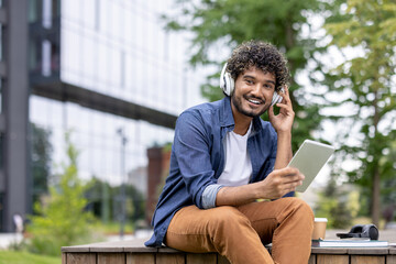 Obraz premium Happy young student man with curly hair wearing headphones, smiling and holding a digital tablet while sitting on a wooden bench outdoors near a modern building on university campus