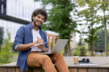 Young mixed-race student with curly hair and earphones sits on a campus bench, smiling while...