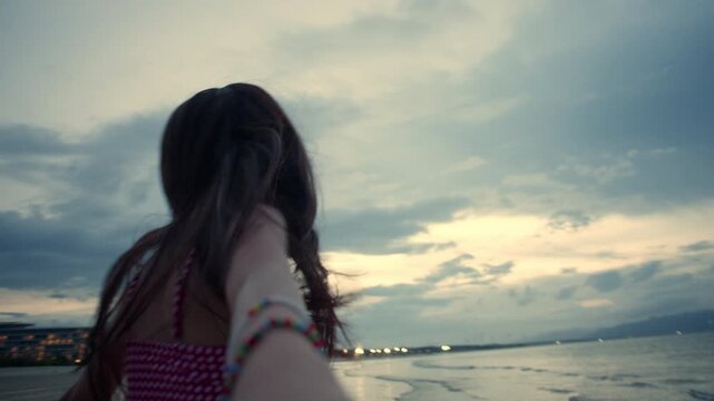 Smiling Asian young woman turns back pulling person along in a dreamy girlfriend POV moment by the seaside at sunset with windblown hair