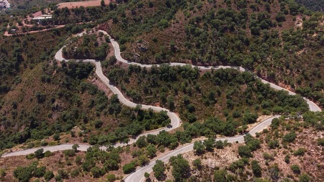 Winding mountain road with sharp hairpin bends cutting through dense green Mediterranean hillside vegetation viewed from above in summer