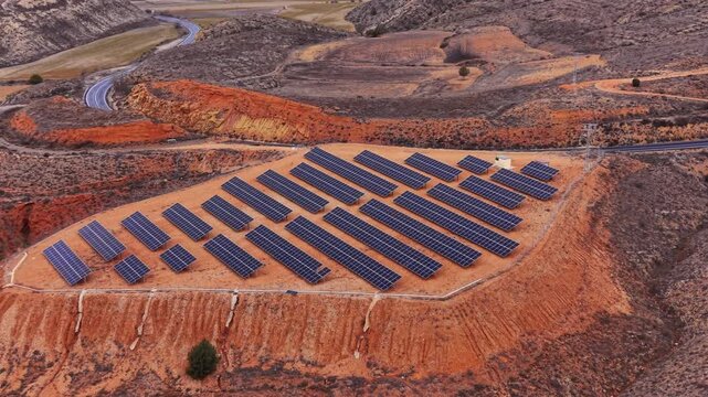 Solar farm with rows of photovoltaic panels on red arid terrain in Spain, surrounded by rocky hills and a winding road cutting through the landscape