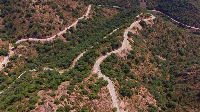 Winding mountain road with sharp curves snaking through green hillsides and shrubland, a white car visible on the switchback asphalt path under clear summer sky