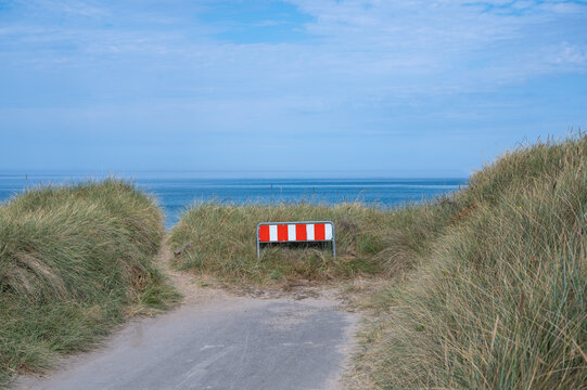 Rot-wei&szlig;es Absperrgitter blockiert einen Weg durch begraste D&uuml;nen zum blauen Meer unter leicht bew&ouml;lktem Himmel an der K&uuml;ste.
