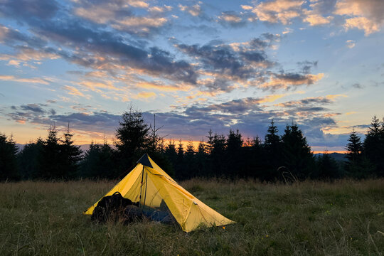 Yellow tourist tent set up in grassy field at sunset, surrounded by trees in evening.