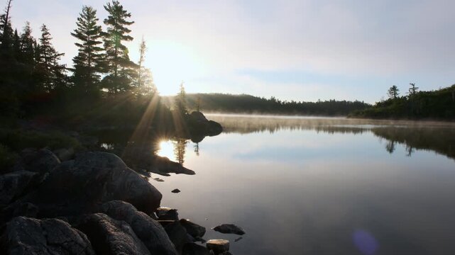 Morning sunlight breaks through pine trees along a rocky shoreline as mist rises off a serene wilderness lake in the Boundary Waters.