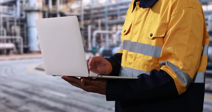 Industrial worker in high visibility yellow uniform using a laptop at an outdoor construction or factory site.