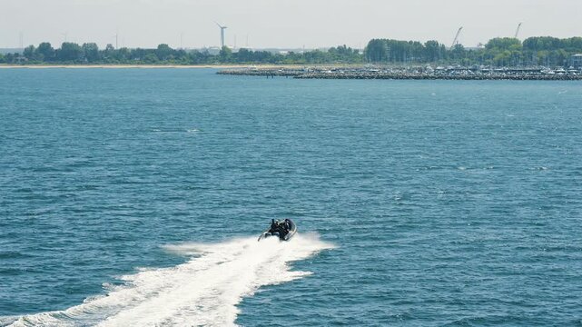 A black RIB speedboat racing toward the coast of Rostock across the Baltic Sea, Germany, throwing up spray with a marina and breakwater in the background on a sunny summer day