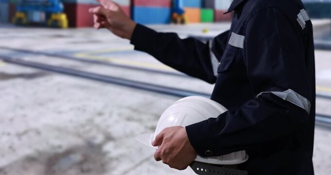 Port worker in dark safety uniform holding a white hard hat and pointing at cargo containers in a busy shipping yard.