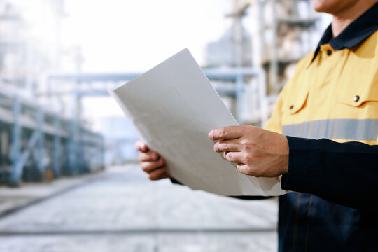 Industrial worker in a yellow safety uniform holds technical documents at a factory site, with blurred machinery and walkways in the background.