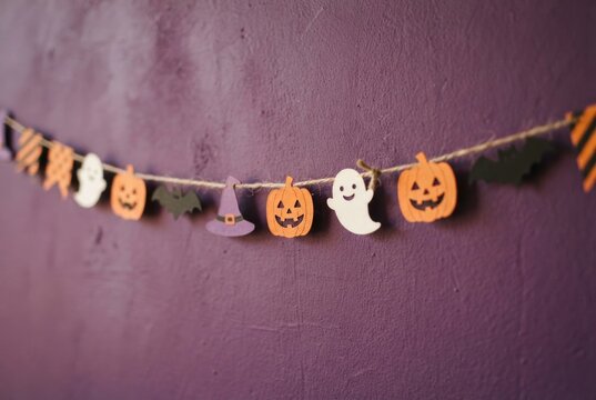 A cheerful Halloween garland with pumpkin, ghost, bat and witch-hat cutouts hangs against a purple wall.
Concept of cute Halloween decorations and festive DIY decor.