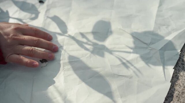 Hand in red sleeve gently touches and smooths creases of crumpled white paper with shadow patterns from nearby foliage. scene is set outdoors on textured stone capturing soft natural light