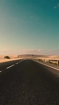 Driver point of view perspective driving on an empty asphalt road that crosses a vast desert landscape with golden sand dunes and a clear turquoise sky, evoking a sense of freedom and travel