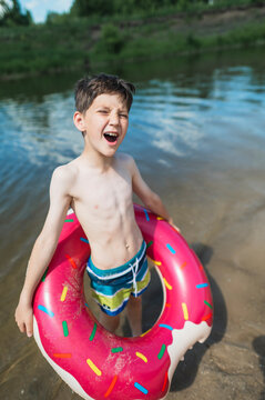 A happy boy stands in the river with a pink donut inflatable ring