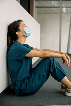 Exhausted nurse in scrubs and mask sitting on hospital floor