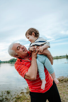 Happy grandfather lifts his baby grandson outdoors by a pond