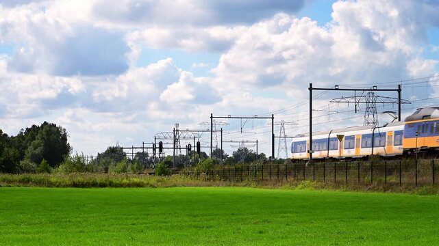 A yellow and blue intercity passenger train of the Nederlandse Spoorwegen NS travels along tracks. Overhead power lines span the sky above. 