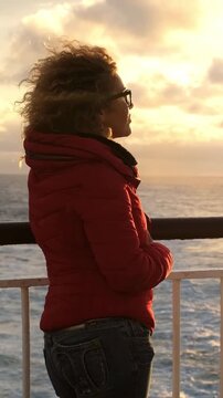 Young woman with curly blonde hair wearing a red jacket, enjoying a scenic ocean sunset from the deck of a moving ferry, her hair blowing in the wind as she gazes at the horizon