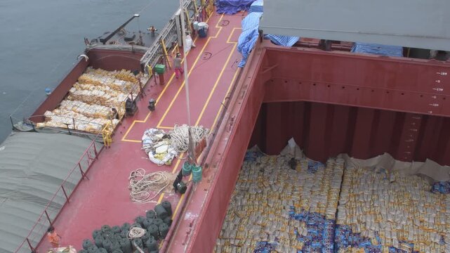 high angle view of a bulk carrier deck and open cargo hold loading bagged goods from an alongside barge with slings, rigging ropes, and manual activities 