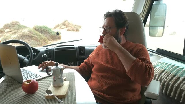 Modern man working as a digital nomad with a laptop inside his camper van, pouring and drinking coffee while enjoying an alternative lifestyle of travel and freedom on the road