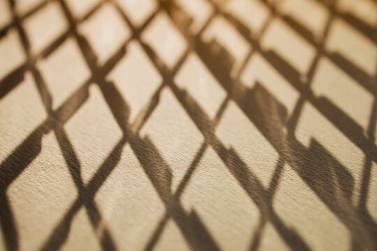Intricate geometric shadows cast by a diamond-patterned metal grate on a smooth surface, creating a visually striking abstract composition in warm tones