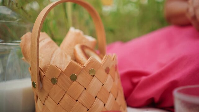 Close up of hand reaching into woven basket to pick out bread and bagels. scene includes fresh baked goods milk in glass carafe pink fabric background soft natural light outdoor greenery