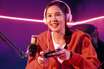 Host channel of smiling beautiful Asian girl streamer with joystick playing online game wearing headphones paste talking with viewers media online. Esport skilled team players in neon room. Stratagem. © InfiniteFlow