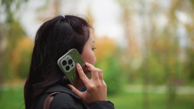 Close up of woman holding smartphone to ear in park setting with greenery and soft lighting. Hand visible phone case distinct background blurred with trees and grass. Natural daylight