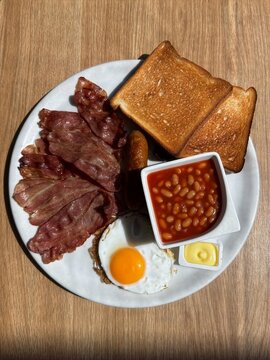Desayuno ingl&eacute;s tradicional completo visto desde arriba sobre una mesa de madera con huevos, bacon, baked beans, tostada y mantequilla