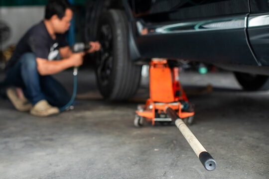 Selective focus to hydraulic floor jack with blurry professional mechanic using a pneumatic wrench to change a wheel in an auto repair shop. Blurred background focuses on the equipment.