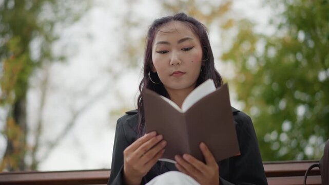 Close up of young woman reading book outdoors on bench with trees in soft focus. Woman holds open brown hardcover wearing dark jacket and white pants scene calm daylight setting