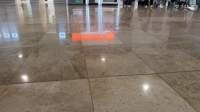Polished stone floor reflects red light projection. People walk through large indoor space. Floor tiles show reflections from overhead lights. Some individuals carry luggage in background