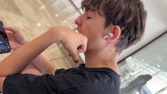 Teenager with earbuds, holding phone, in indoor space. Hand near mouth, looking at screen. Light reflections show glass surfaces. Casual posture, relaxed setting