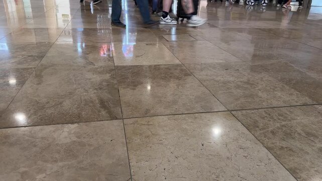 Large polished stone tiles cover ground. People walk past in background. Reflections appear on shiny floor surface. Footage shows indoor public space. Floor tiles show natural patterns