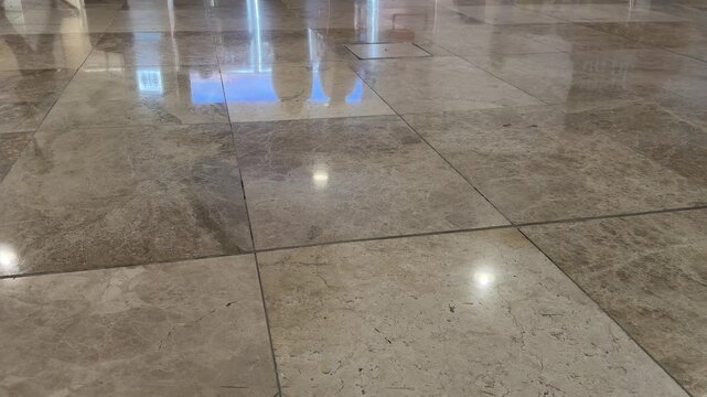 Person walks on polished beige stone floor. Shoes step forward in slow motion. Reflections show bright light above. Floor tiles shine with natural texture. Person's legs and feet appear briefly