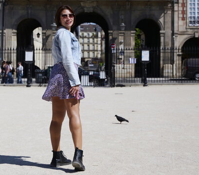 An Asian woman wearing a light denim jacket, a purple floral mini skirt, and black boots stands smiling looking back over her shoulder in a paved courtyard near arched stone architecture