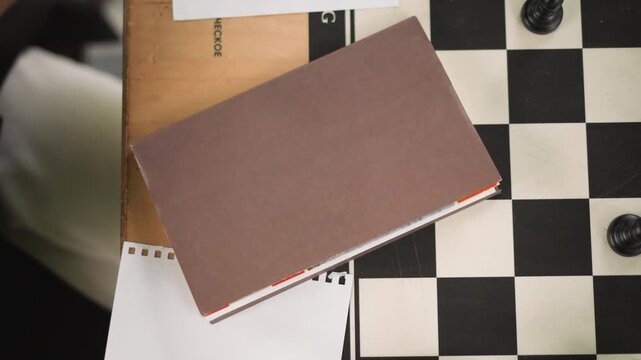 Close up of brown notebook on checkered surface with chess pieces wooden board and paper. Hand briefly visible interacting near objects in soft lighting. Scene is stationary with clear focus
