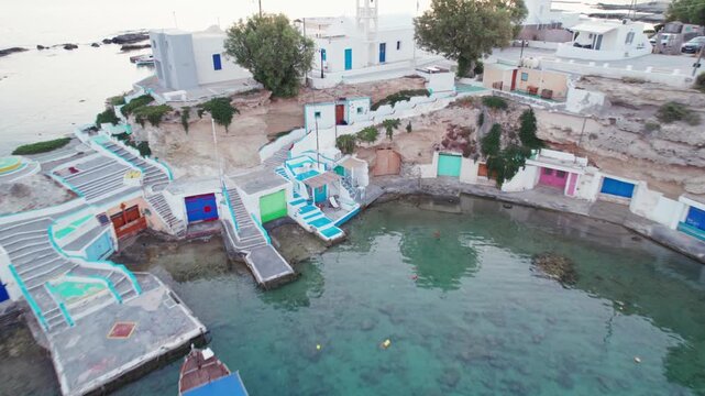 Aerial drone shot of vibrant syrmata boathouses carved into the rocky shoreline at Mandrakia village, Milos, Greece, featuring painted stairs and calm clear waters