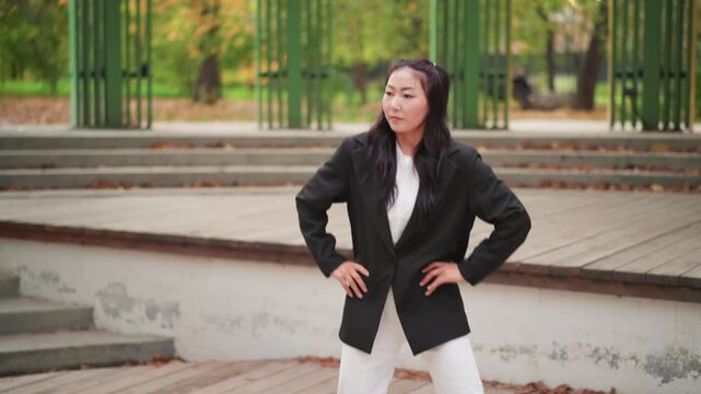 Close up of young woman in professional attire performing dance movements on outdoor wooden platform with green metal structures and trees visible in background. Her hands on hips pose